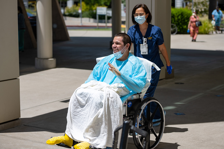 Gabe Gonzalez sits in wheelchair wearing blue hospital gown while a nurse in scrubs pushes him outside