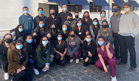 A group of 27 volunteers wearing face masks and many in scrubs, with some standing and others squatting, pose for a photo