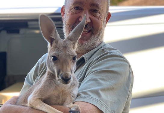 A smiling man with white hair and a beard holds a baby kangaroo in his arms