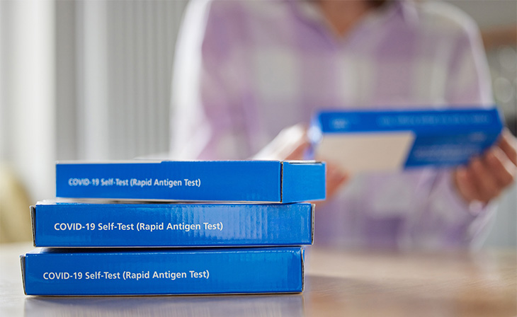 A blurry person is seen in the background holding a blue COVID-19. Three blue boxes are in the foreground and have white lettering with the words COVID-19 Self-Test (Rapid Antigen Test).