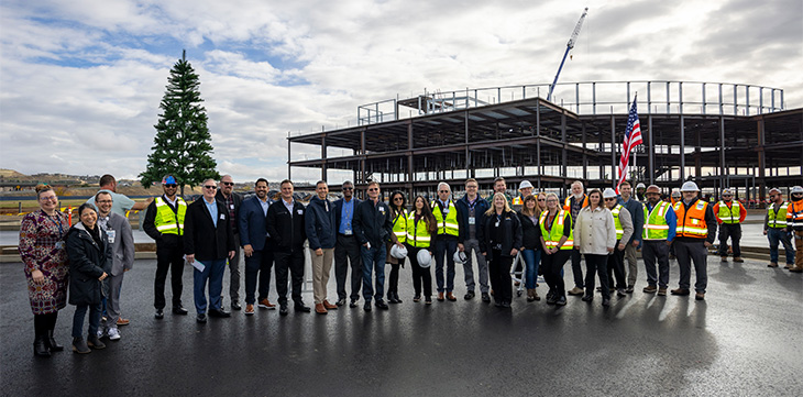 A large group of people stands outside in front of a large steel beam for a group photo.