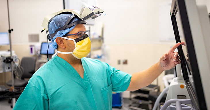A surgeon wearing green scrubs, a cap and an augmented reality headset stands works with a computer screen in an operating room. 