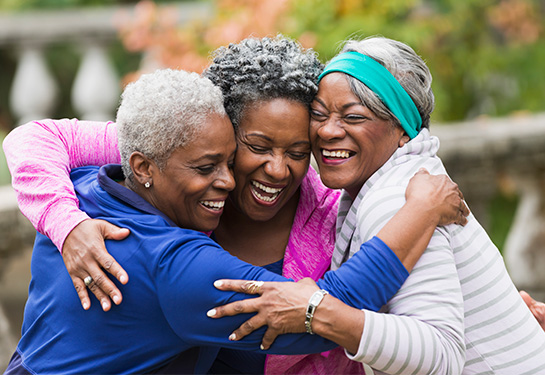 Three older smiling African American women in an outdoor park embrace in a group hug.