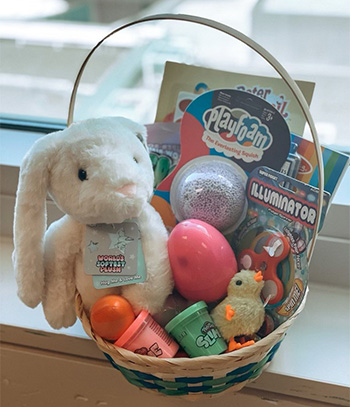 Basket on a windowsill is filled with a white stuffed rabbit, containers of slime, plastic eggs and a small stuffed chick. 