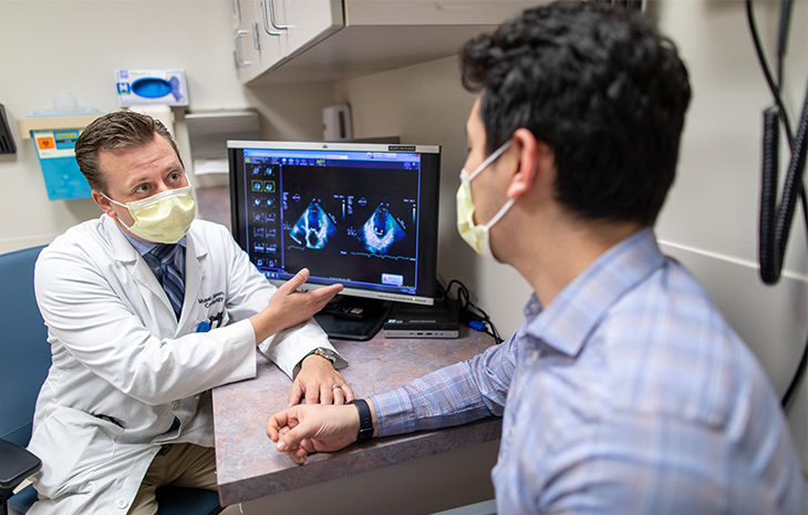 Doctor in a white lab coat and mask over his face speaks with a patient about an ultrasound image on a computer screen.