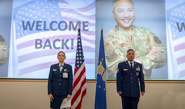 Two people dressed in Air Force uniforms stand attention in front of a large screen that reads “Welcome Back”