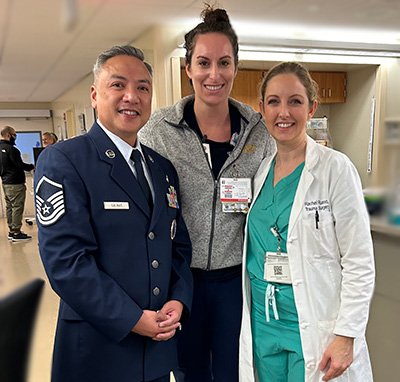 Three people, one in hospital scrubs and white coat, one in Air Force uniform and one in black pants and gray jacket, pose in a hospital environment. 