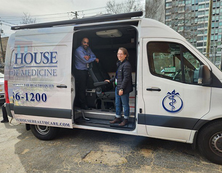 Two people stand inside a white van with its sliding door open and the words “House of Medicine” on the side panel
