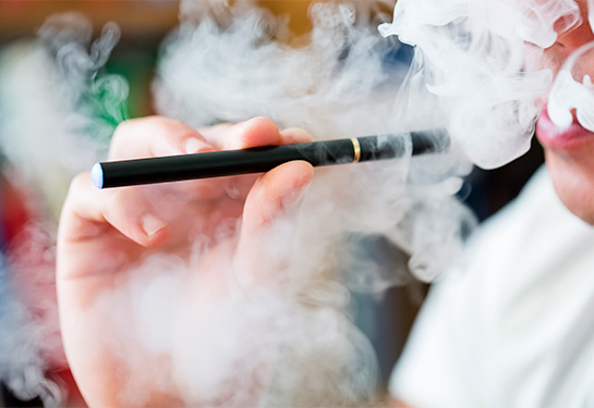 A hand holds a black e-cigarette surrounded by a cloud of white smoke