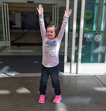 Smiling child with pink shoes on with her hands in the air standing outside a building. 