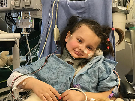 Smiling child in a blue hospital gown with neck brace sitting in a chair.