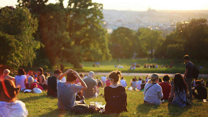 Dozens of people sitting on a grassy hillside overlooking a city sunset