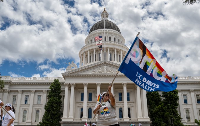 UC Davis Health Pride event at state capital in Sacramento, participant waves Pride flag with capital building in background.