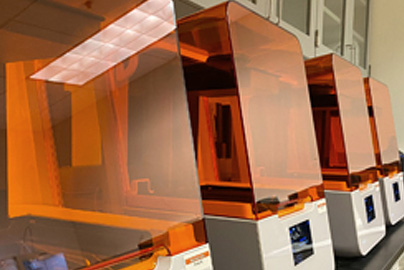 Four 3D printers with orange covers sitting on a table in a row