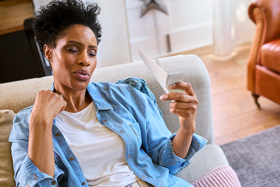Woman sitting on a couch experiencing a hot flash while holding a handheld fan.