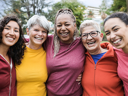 Five diverse women smiling together outdoors with their arms around one another.
