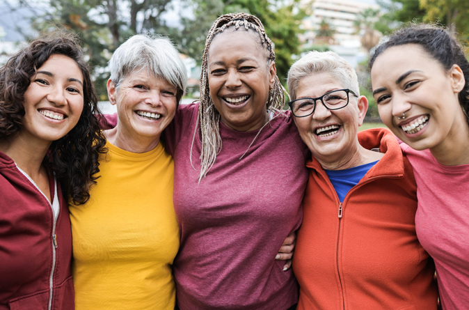Five diverse women smiling together outdoors with their arms around one another.