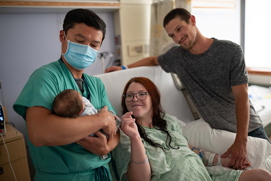 Kayla in a hospital bed with her newborn being held by health care worker next to her