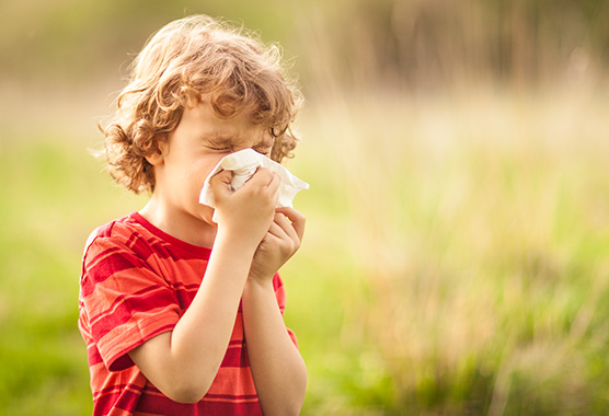 A boy sitting in a field of grass holding a tissue and sneezing.
