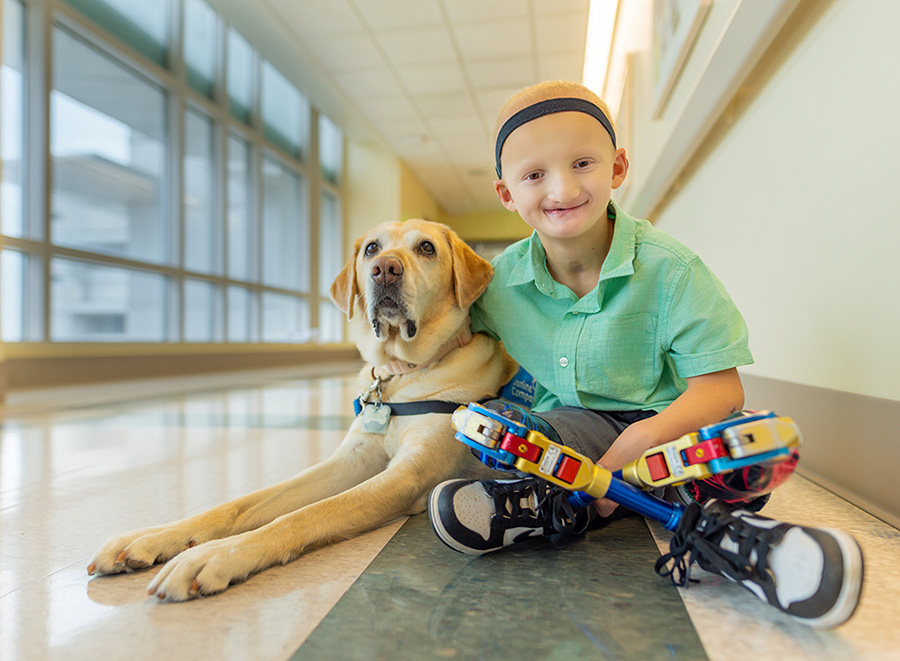 : A photo of a young child sitting with a hospital facility dog in a light filled hallway in the children’s hospital.