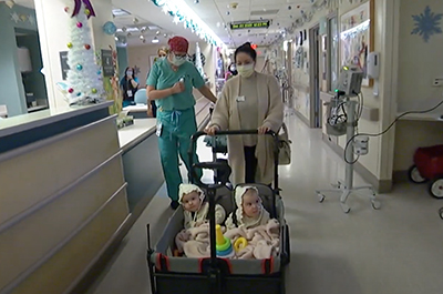 Separated twins in a hospital hallway being pushed in a wheeled cart. 