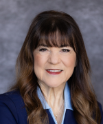 A person with long dark hair and a dark jacket over blue shirt smiles.