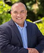A person in a dark blue suit and shirt smiles at the camera with arms crossed.