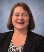 Smiling person in a dark blazer and patterned blouse against a blue-gray background.
