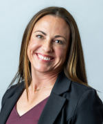 Smiling person with straight shoulder-length brown hair wearing a dark blazer over a maroon top against a plain light blue background.