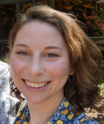 Smiling person outdoors with brown hair and dangling earrings, wearing a blue patterned top.