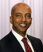 Smiling bald person in a black suit, white shirt, and red patterned tie, posed against light gray curtains.