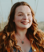 A person with long auburn hair and a necklace in an outdoor setting.