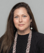 A person with long brown hair, wearing a dark top and a patterned, beaded necklace, against a plain light background.
