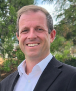 A smiling person with short blond hair, wearing a suite and white shirt, against an outdoors backgound.