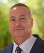 A person in a suit and tie, facing the camera with a neutral expression, set against a softly blurred green outdoor background.