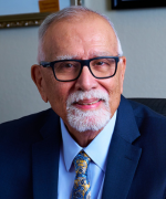 A person in a blue suit and patterned tie, wearing glasses, smiling toward the camera in an office.