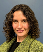 Portrait of a person with dark curly hair wearing a green textured jacket, facing the camera against a plain dark background.