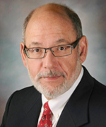 A person in a dark suit, white shirt, and red tie, wearing rectangular glasses and a beard, against a plain gray background.
