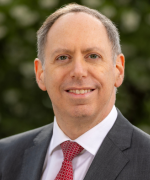 Smiling person in a dark suit and white shirt with a red patterned tie.