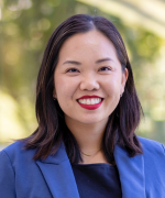 Smiling person with shoulder-length dark hair wearing a navy blazer and red lipstick, outdoors with green trees behind her.