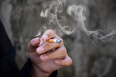 A hand holds a lit cigarette, releasing white smoke into the air against a blurred outdoor background.