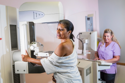 A Black woman step toward a mammogram machine as a smiling woman nurse stands nearby. 