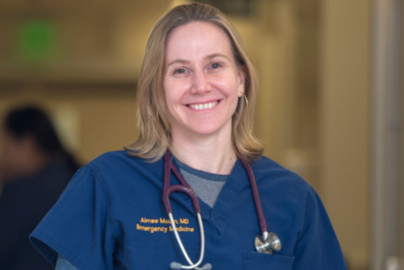A person wearing blue medical scrubs with a stethoscope stands in sharp focus in a healthcare setting.
