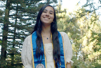 A woman with dark hair and a white shirt smiles in front of trees.