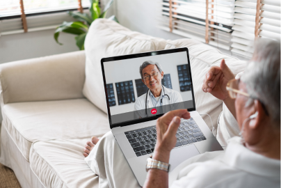 An older man sits on a couch with a laptop on a video call with a doctor. 