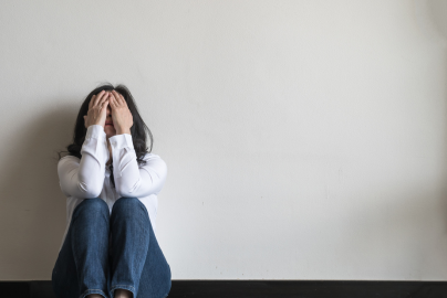 A woman in a white shirt sits on the ground against a wall with hands over her face.