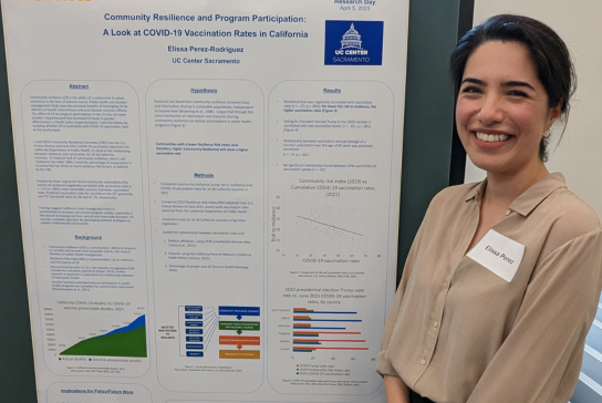 A woman with dark hair in a light colored shirt smiles in front of her research poster. 