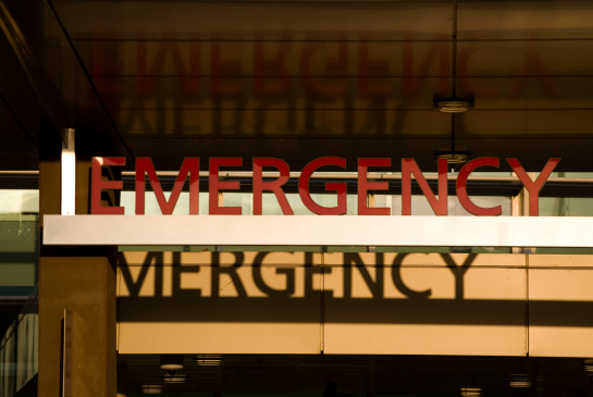 An "Emergency" sign on a hospital is above a door and reflecting off a window. 