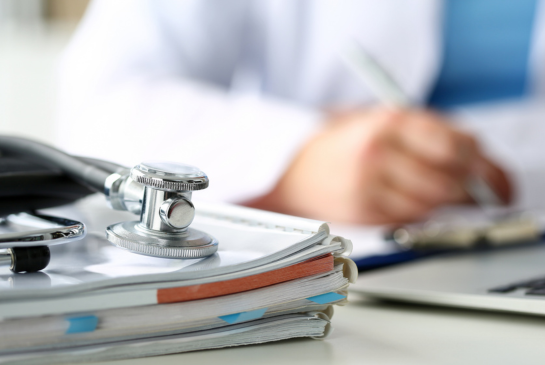 A stethoscope sits on top of a pile of reports as a doctor writes on a desk in the background.