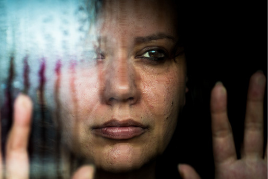 A woman with dark hair looks sad through a rainy window. 